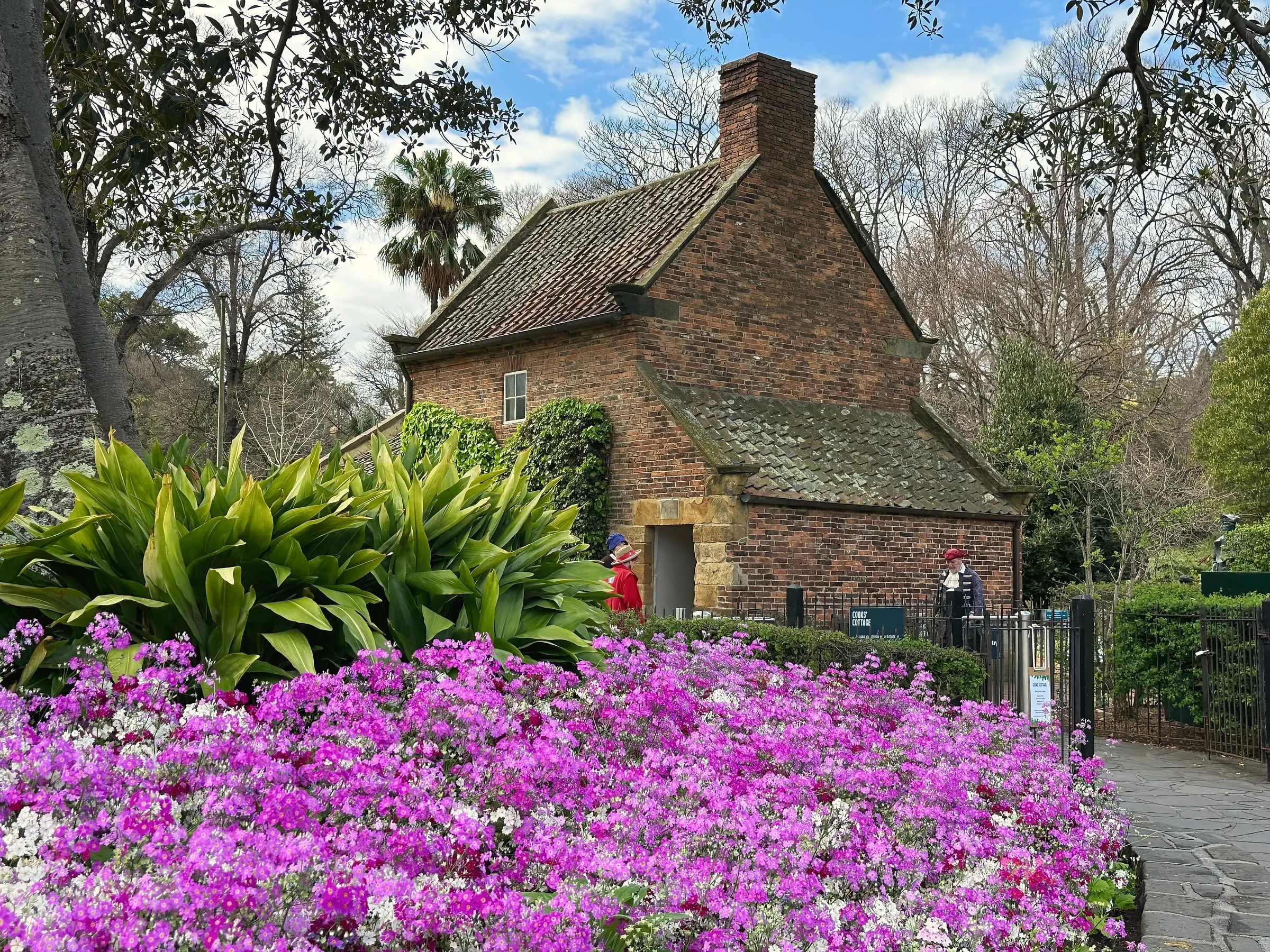 An old brick cottage with a tall chimney behind a bed of purple and white flowers