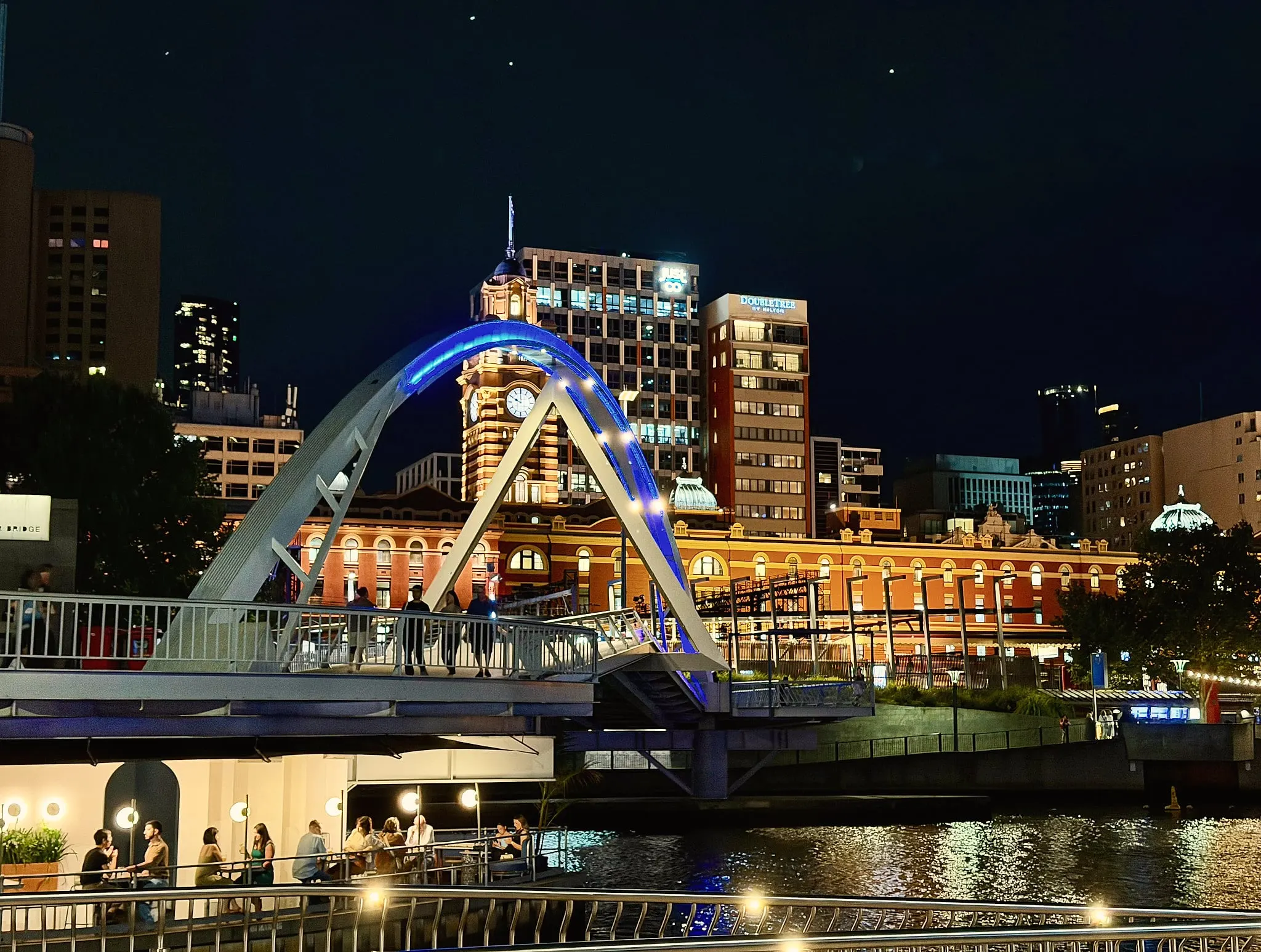 An arched footbridge lit blue at night over the Yarra, with Flinders Street Station glowing behind