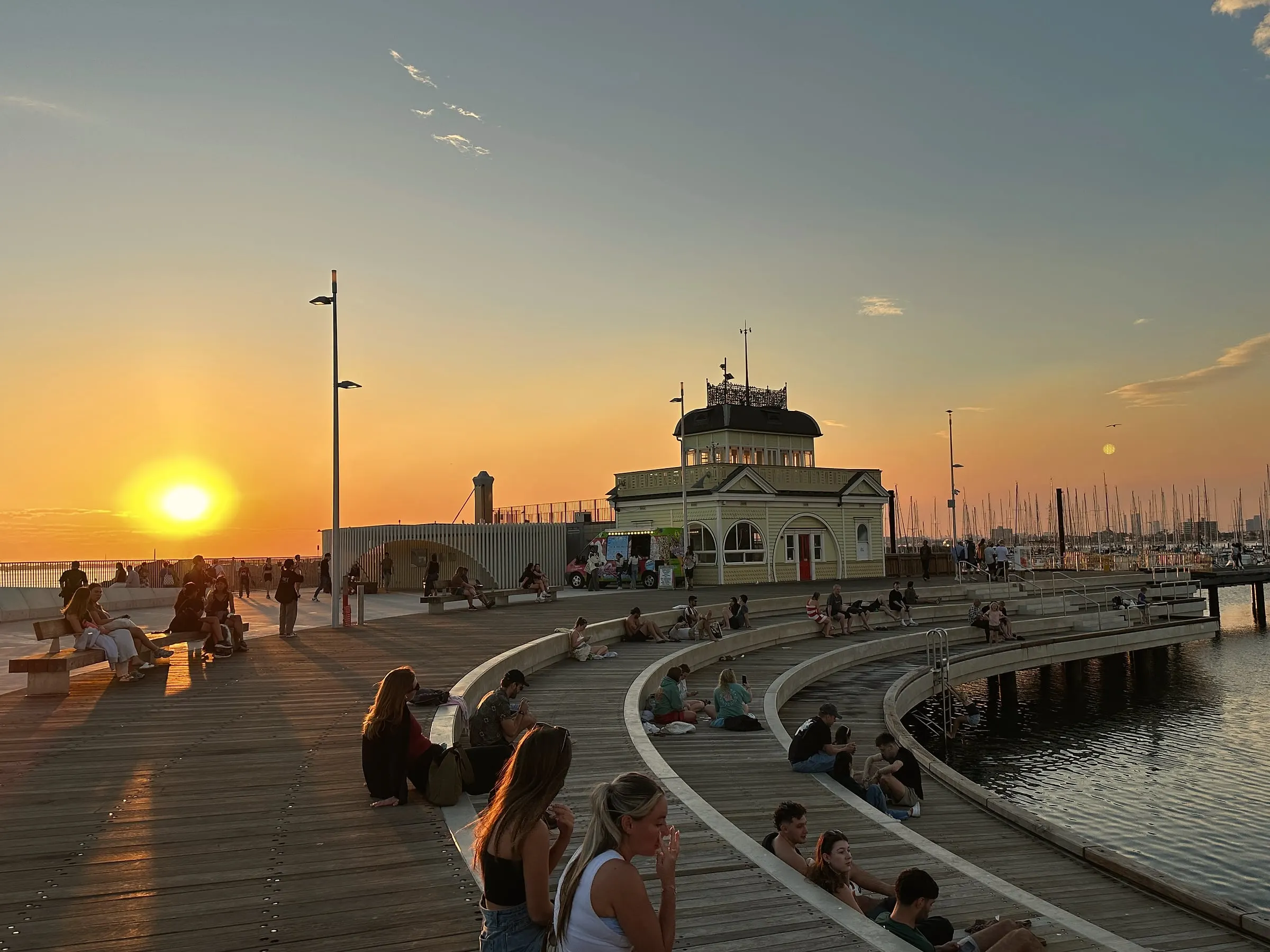 People sitting on curved timber steps on a pier, watching the sun set beside a historic kiosk