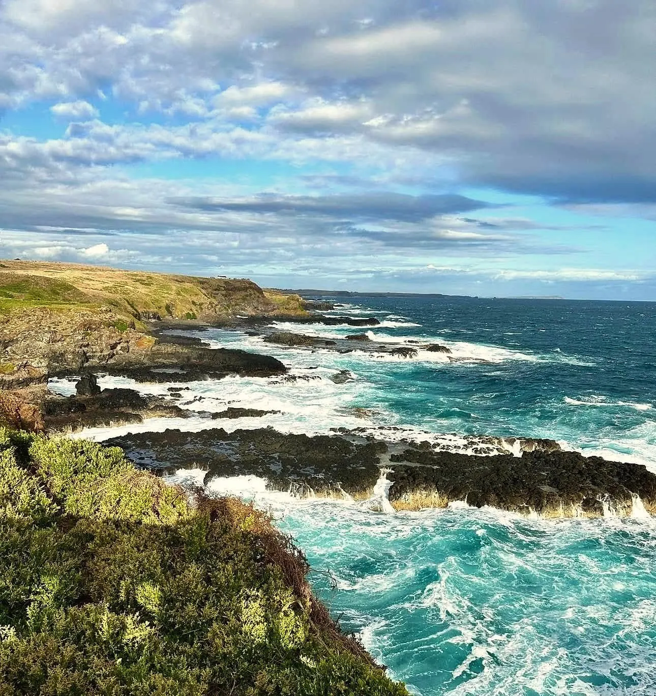 Waves breaking white over dark rocks below a green clifftop, with the coastline stretching away under a cloudy sky