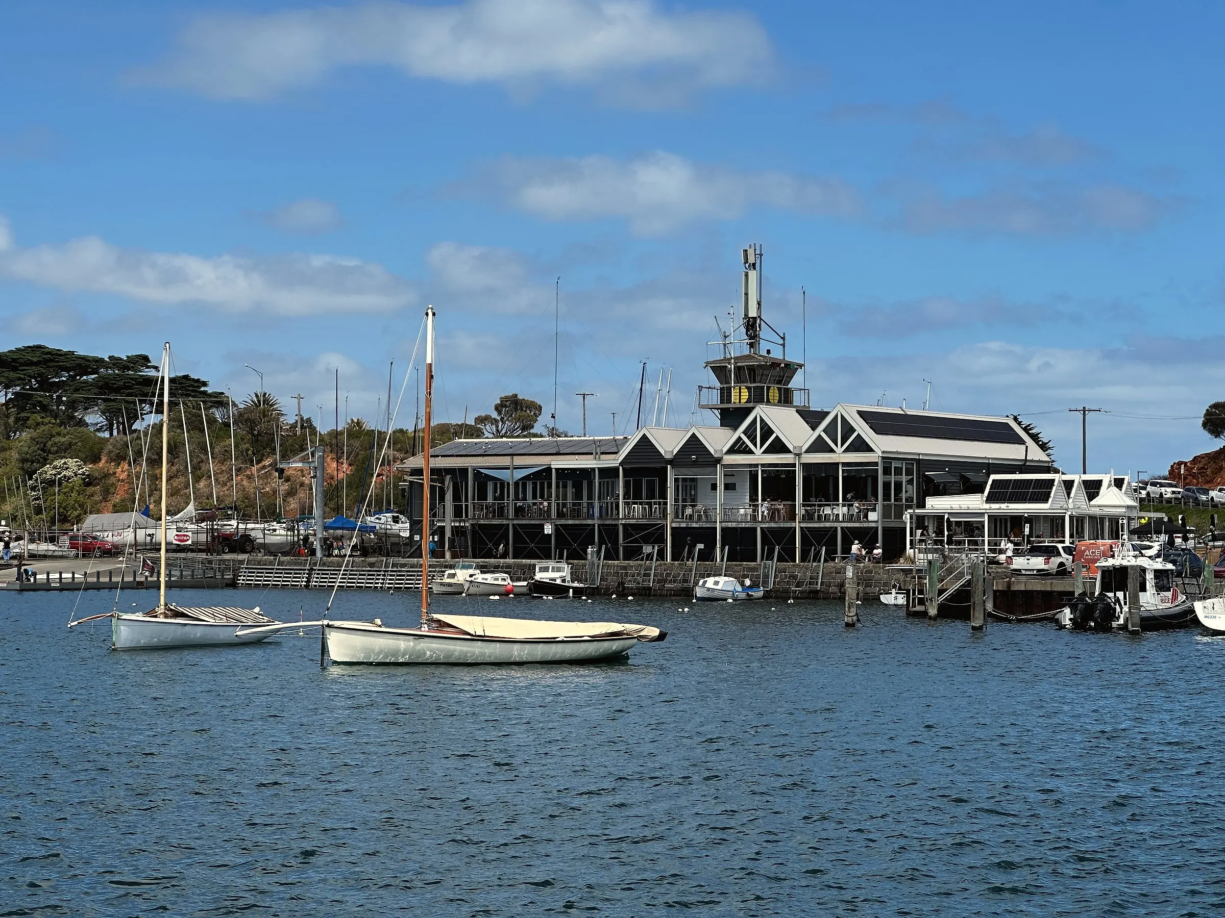 Small sailing boats moored in a calm harbour in front of a yacht club building with a tower