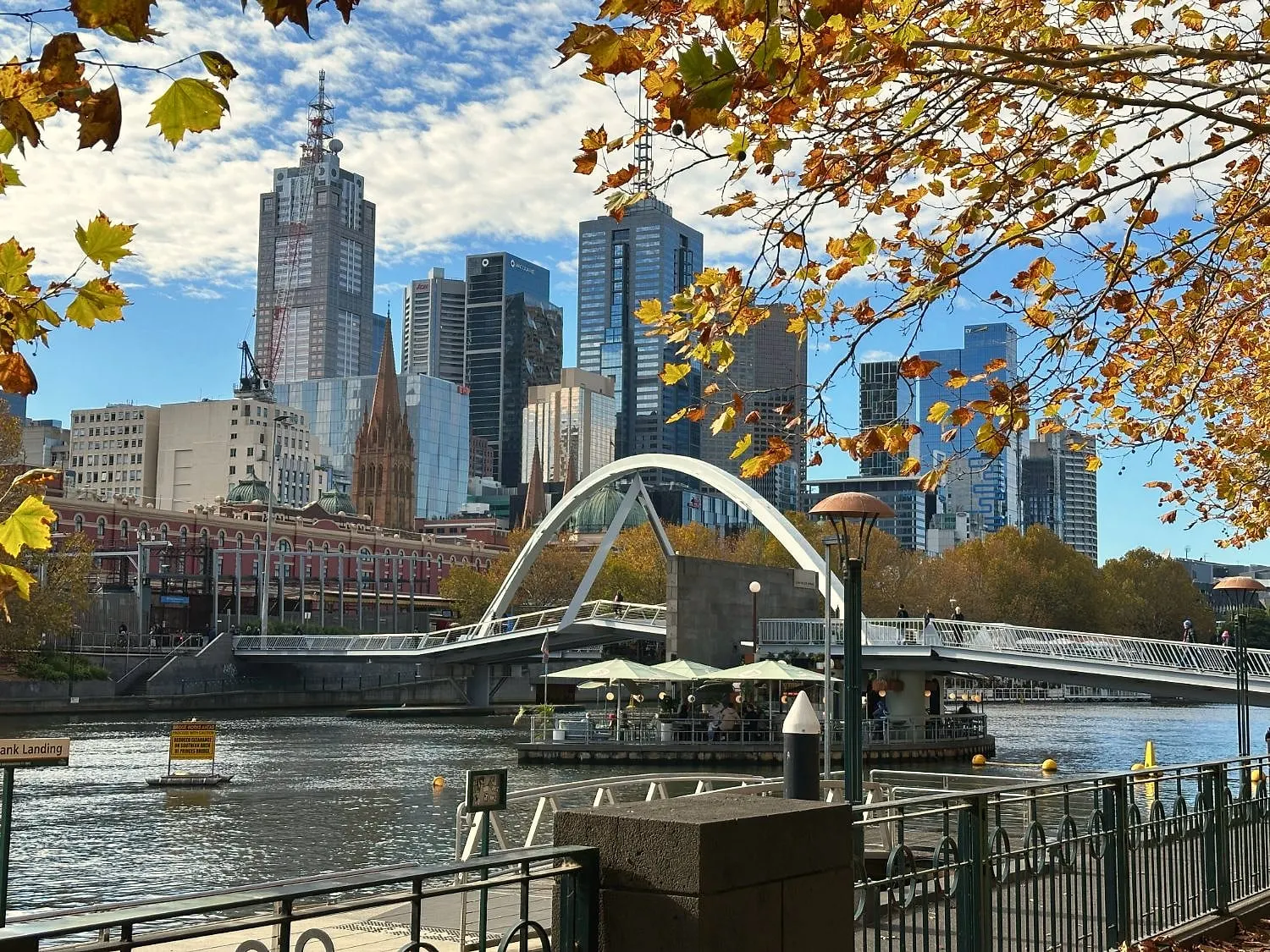 An arched footbridge over the Yarra with city towers behind, framed by golden autumn leaves