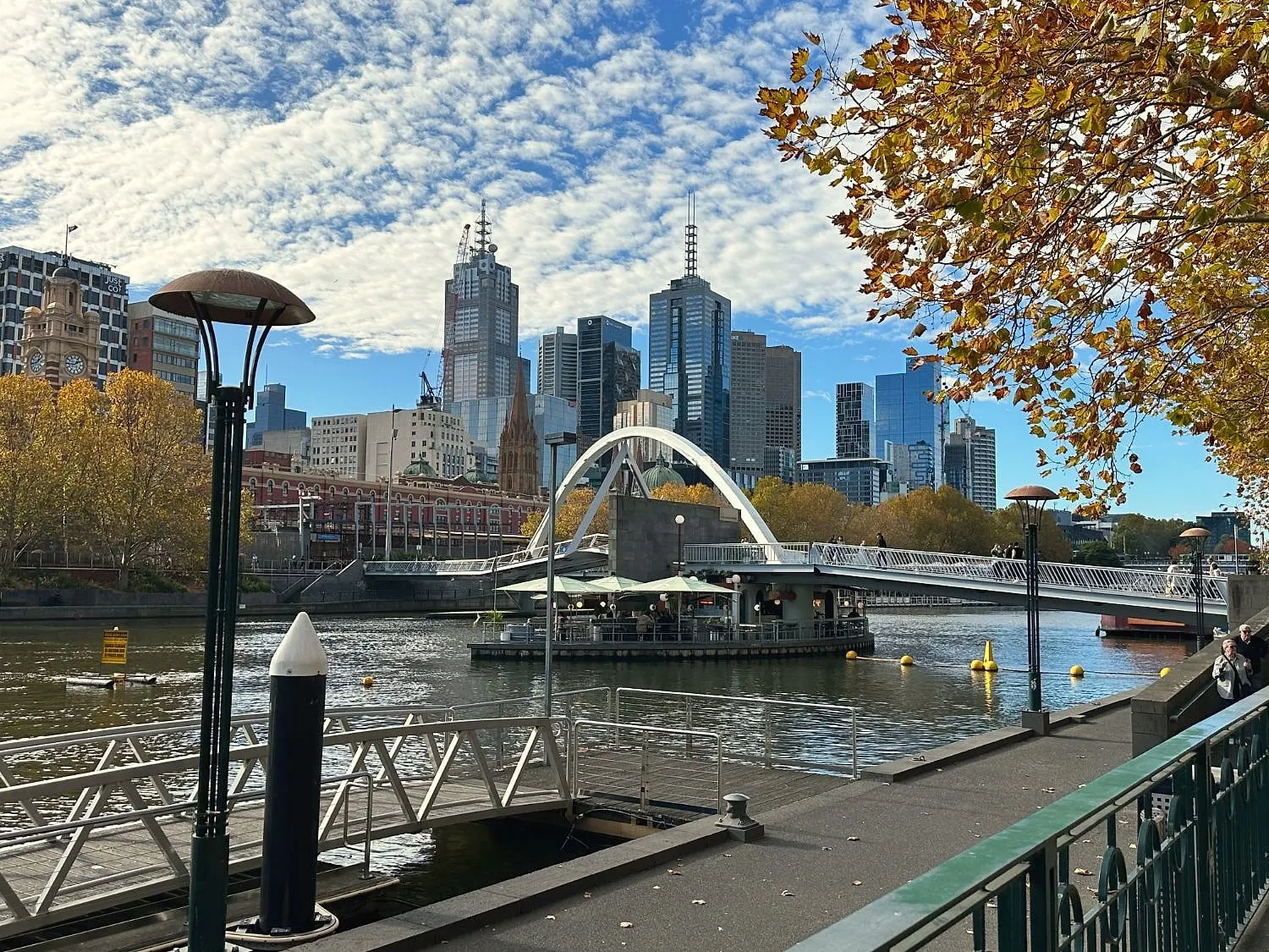 The Yarra River from the Southbank promenade, with an arched footbridge, Flinders Street Station and city towers beneath golden autumn trees